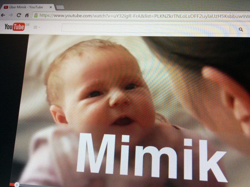 Ein Screenshot des YouTube-Kanals "Singale-des-Babys" Ein-Video-Lexikon-auf-DVD-und-YouTube zeigt ein Baby von vorne gehlaten von seiner Mutter und den Schriftzug "Mimik"