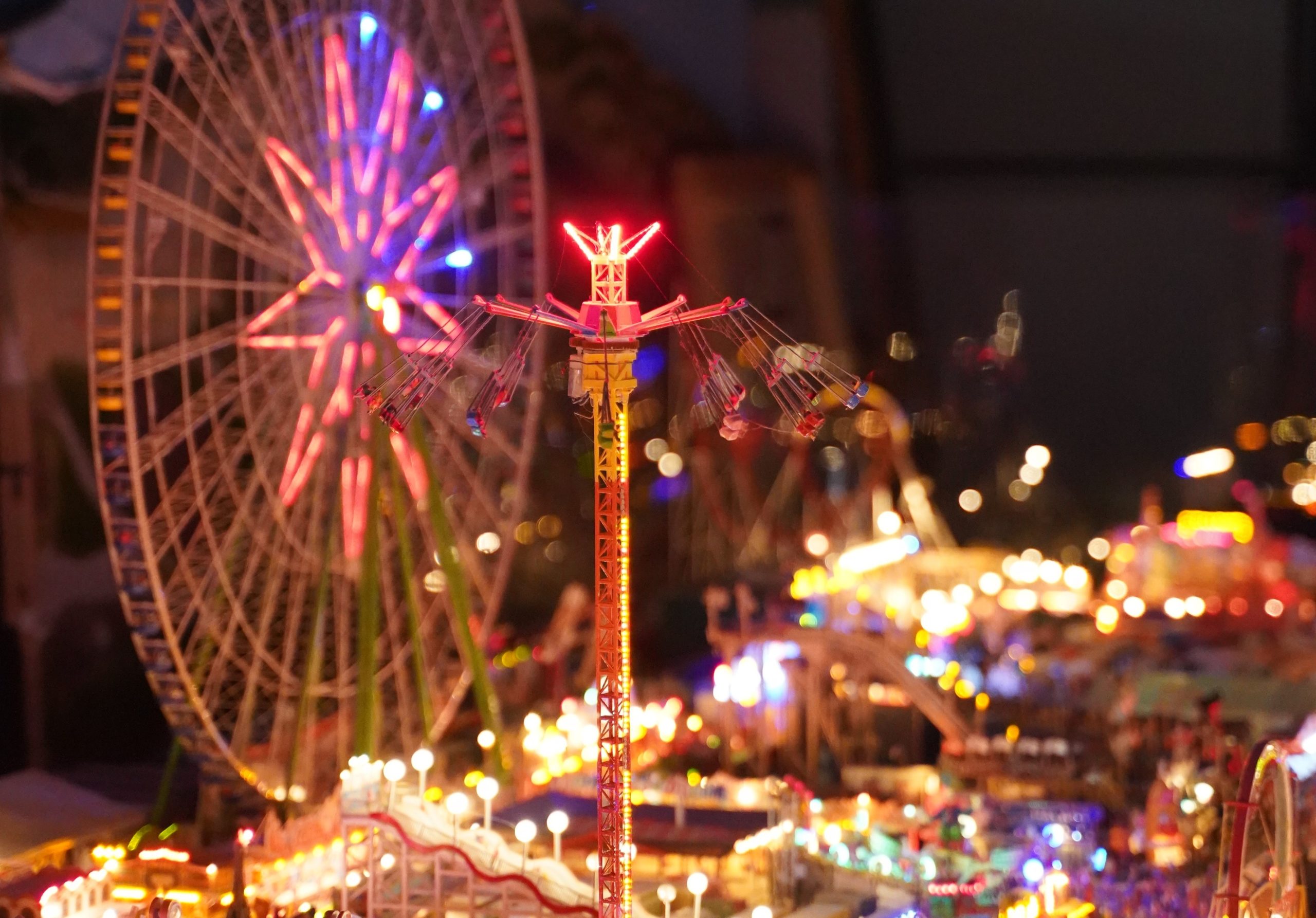 Die Wiesn bei Nachtstimmung, bunte Lichter erscheinen von den Fahrgeschäften und hellen die Atmosphäre auf. Im Vordergrund sieht man einen freien Fall Tower (Fahrgeschäft).