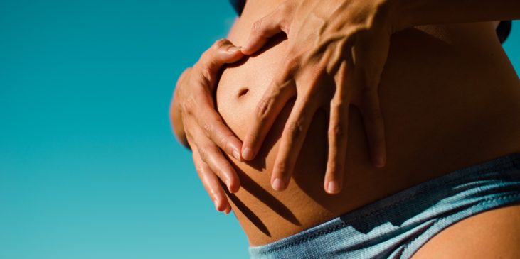 Der nackte Bauch einer Schwangeren Frau im Bikin von zwei Händen schützend gehalten
