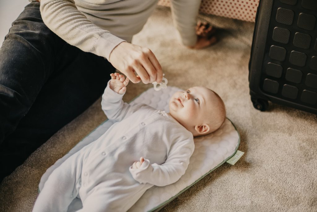 Ein Elternteil sitzt neben einem Baby auf dem Boden und hält dem Baby den Schnuller hin. Das Baby greift danach und lacht das Elternteil an. Von dem Elternteil sieht man nur die Beine und einen teil des Oberkörpers.