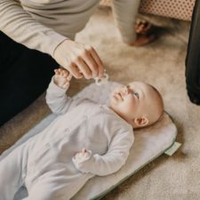 Ein Elternteil sitzt neben einem Baby auf dem Boden und hält dem Baby den Schnuller hin. Das Baby greift danach und lacht das Elternteil an. Von dem Elternteil sieht man nur die Beine und einen teil des Oberkörpers.