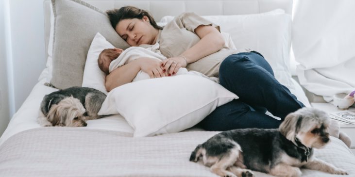 Eine Frau liegt auf dem Sofa und scheint zu schlafen. Sie trägt T-Shirt und Jeans. In Ihren Armen und auf ein Kissen gebettet schläft ein Baby.