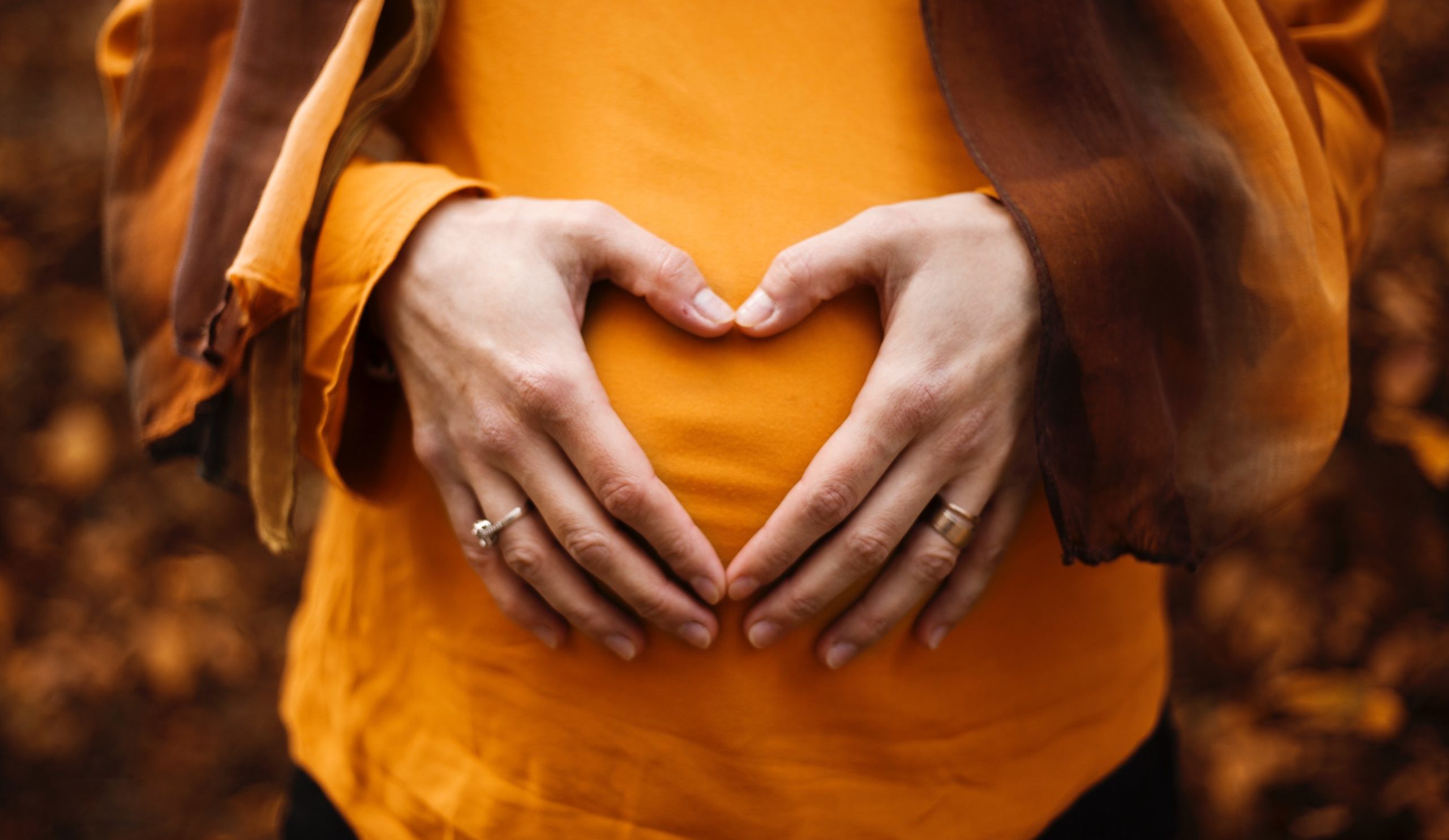 Eine schwangere Frau hält mit ihren beiden Händen sanft ihren Babybauch und formt dabei ein Herz mit ihren Fingern. 