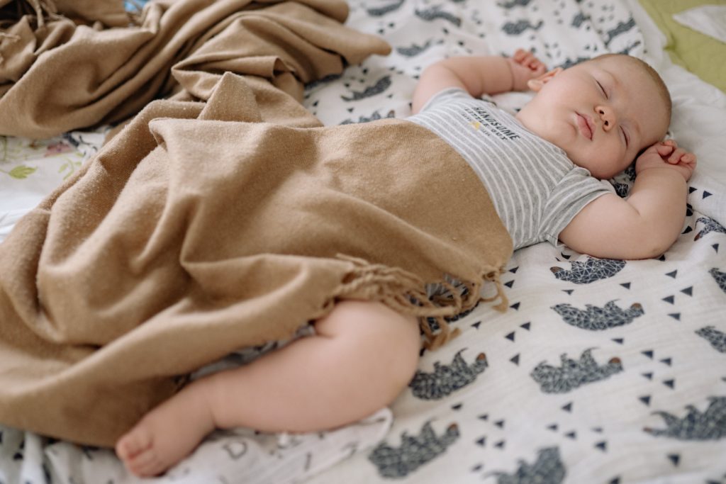 Ein etwa 5-6 Monate altes Baby liegt auf einer Decke mit Elefantenmuster. Es schläft und hat dabei die Arme neben dem Kopf abgelegt. Ein Bein und ein Teil des Oberkörpers ist mit einer braunen Decke zugedeckt.