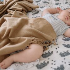 Ein etwa 5-6 Monate altes Baby liegt auf einer Decke mit Elefantenmuster. Es schläft und hat dabei die Arme neben dem Kopf abgelegt. Ein Bein und ein Teil des Oberkörpers ist mit einer braunen Decke zugedeckt.