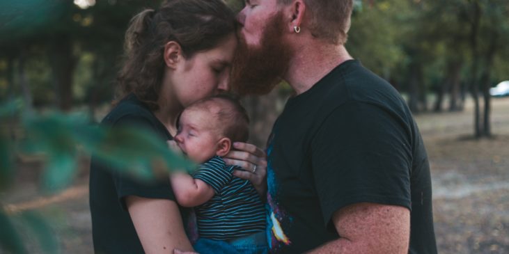 Ein Mann mit kurzen Haaren un dlangem, sehr dichten Barte küsste seine Frau, die ihm gegenüber steht, auf die Stirn und berührt ihren Oberarm. Die Frau halt ein Baby im Arm