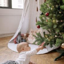 Ein etwa 6 Monate altes Kind liegt in Bauchlage auf dem Boden. Vor ihm ist ein geschmückter Tannenbaum. Das Kind greift mit der rechten Hand nach den Zweigen. Im Hintergrund sieht man ein Zelt mit Spielsachen und ein Fenster.