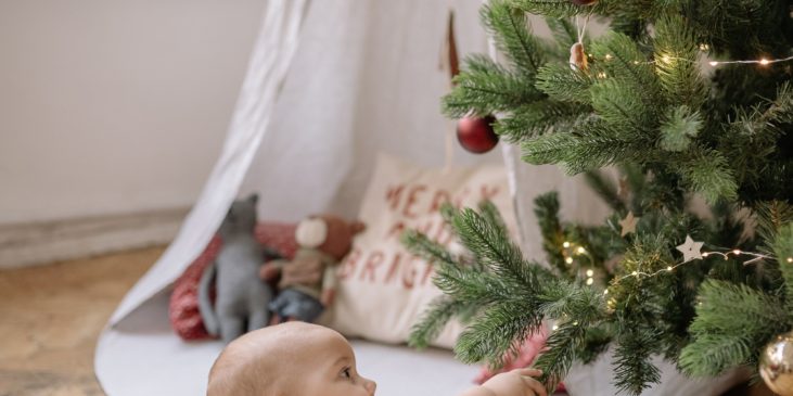 Ein etwa 6 Monate altes Kind liegt in Bauchlage auf dem Boden. Vor ihm ist ein geschmückter Tannenbaum. Das Kind greift mit der rechten Hand nach den Zweigen. Im Hintergrund sieht man ein Zelt mit Spielsachen und ein Fenster.