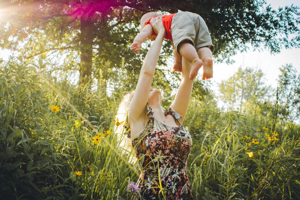 Eine Frau, die ein buntes Sommerkleid mit Trägern anhat, steht in einer Wiese mit hohem Gras. An den ausgestreckten Armen hält sie ein etwa 6 Monate altes Kind in die Höhe und guckt es an. 