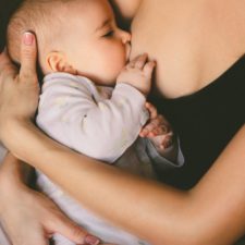 Man sieht den Ausschnitt des Oberkörpers einer Frau, an deren rechter Brust ein Kind gestillt wird. Das Kind hat eine Hand an der Brust. Die Mutter umfasst das Kind zärtlich mit beiden Armen und hält es.