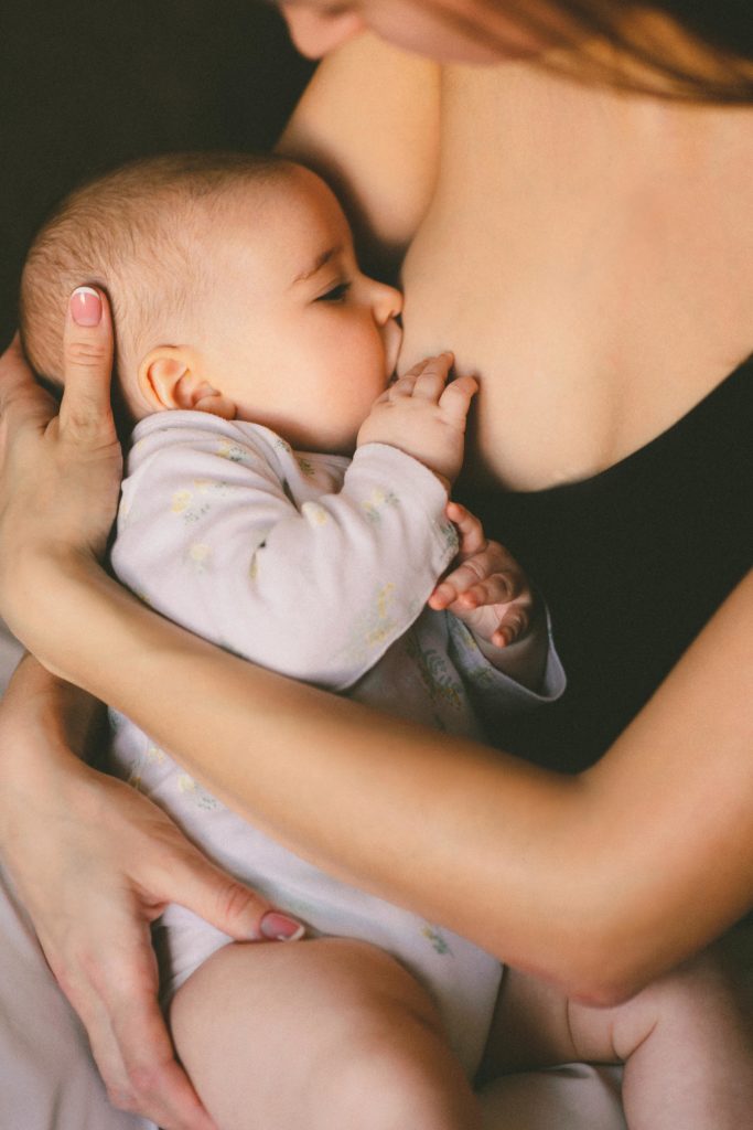 Man sieht den Ausschnitt des Oberkörpers einer Frau, an deren rechter Brust ein Kind gestillt wird. Das Kind hat eine Hand an der Brust. Die Mutter umfasst das Kind zärtlich mit beiden Armen und hält es. 