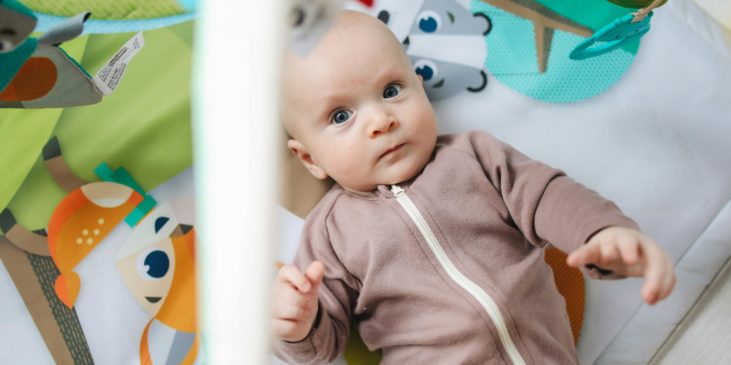 Ein Baby mit braunem Strampelanzug liegt unter einem Spielebogen auf einer Decke mit Tierbildern und guckt ernst in die Kamera