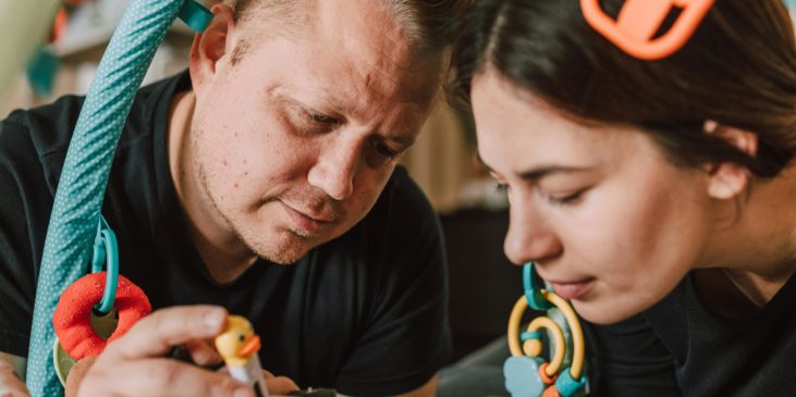 Ein Mann und eine Frau beugen sich über ein Kind. Alle gemeinsam liegen unter einem Spielebogen. Die Frau legt dem Kind zärtlich die Hand an den Kopf. Der Mann hält ein Spielzeug in der Hand.