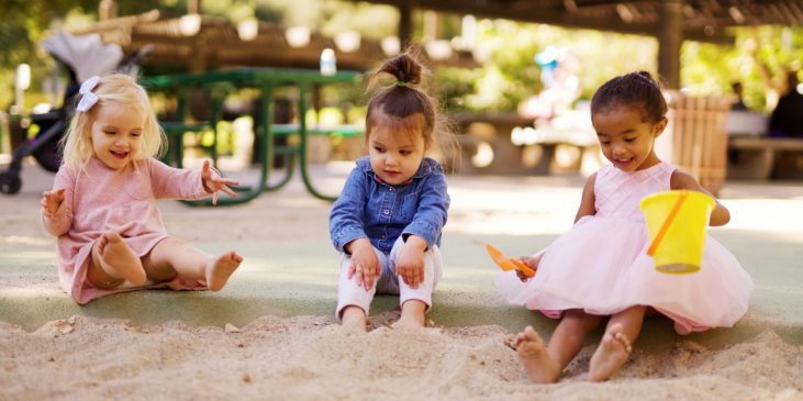 Drei kleine Mädchen sitzen in einem Sandkasten und halten ihre nackten Füße in den Sand. Ein Mädchen ist blond, ein Mädchen hat braune Haare und ein Mädchen hat etwas dunklere Haut. Alle haben lange Haare und lachen fröhlich.