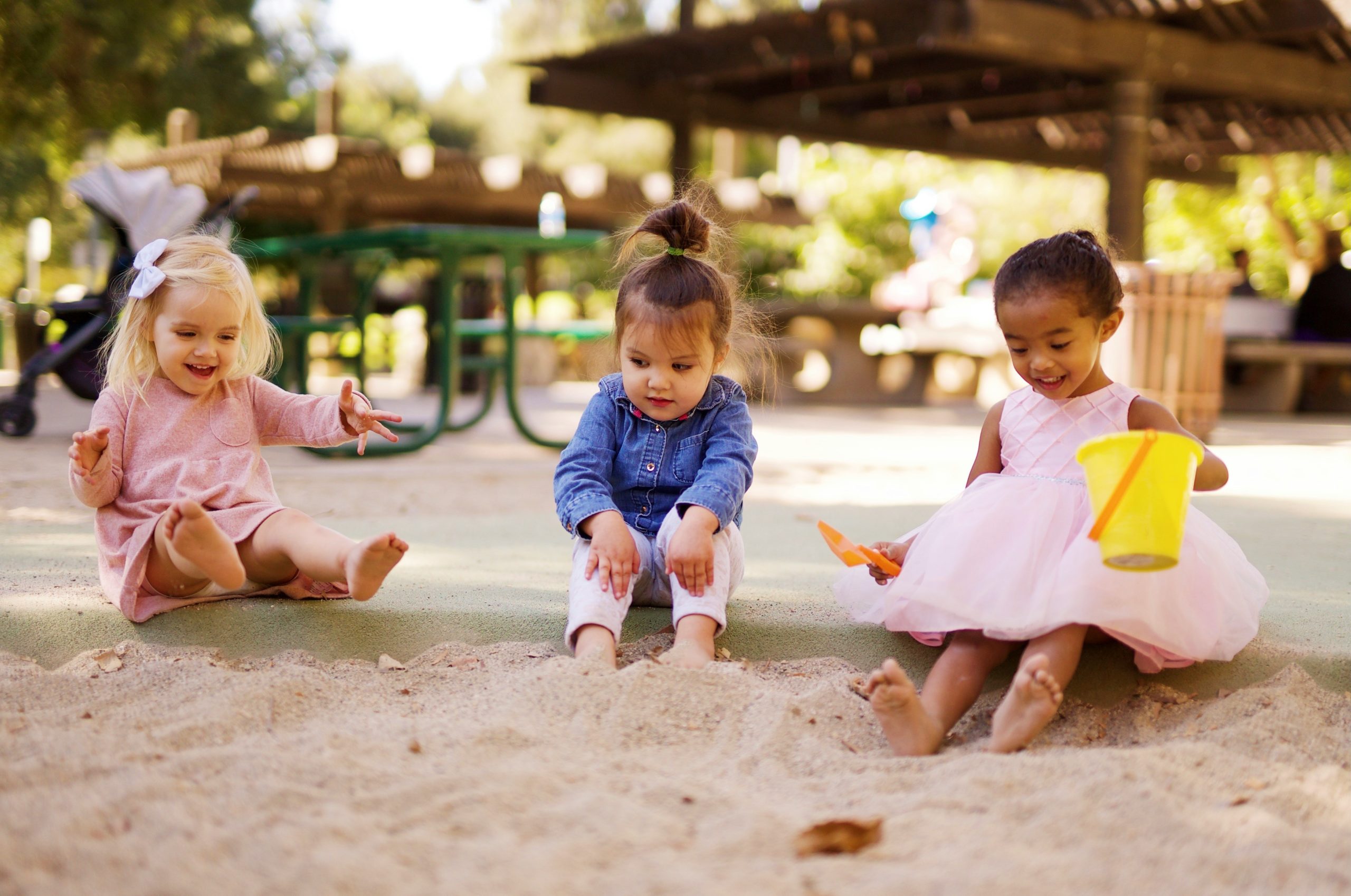 Drei kleine Mädchen sitzen in einem Sandkasten und halten ihre nackten Füße in den Sand. Ein Mädchen ist blond, ein Mädchen hat braune Haare und ein Mädchen hat etwas dunklere Haut. Alle haben lange Haare und lachen fröhlich.